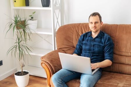 Work remotely, work at home. Young positive man sitting on a sofa with a laptop and looking at the cameraの写真素材