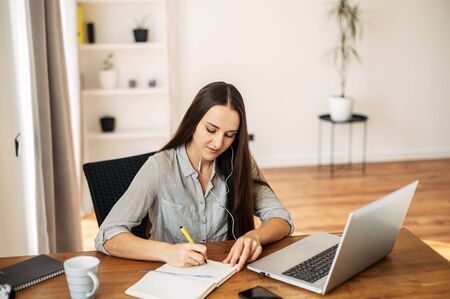 Work in the office, work at home. A young woman writes something in a notebook with a pen, headphones in her ears and a laptop on the tableの写真素材