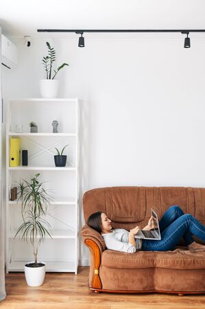 Home leisure. A young woman is lying at home on the sofa and using a laptop. Online shopping, web surfing, social networks, watching videoの写真素材