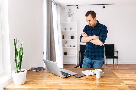 Male office worker, freelancer looking for a solution to a problem. He stands in front of a laptop with his arms crossedの写真素材
