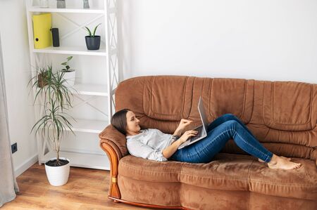 Home leisure. A young woman is lying at home on the sofa and using a laptop. Online shopping, web surfing, social networks, watching videoの写真素材
