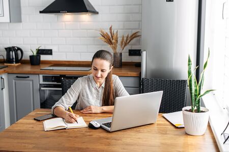 Online training. Girl student watching a lecture, webinar using a laptop. She sits in the kitchen at the table and writes information with a pen in a notebook.の写真素材