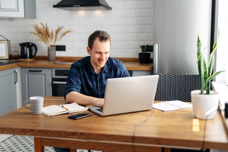 Work remotely, office at home. Pleasant young man uses a laptop for work. He is sitting in the kitchen at home, looking at the screen and smilingの写真素材