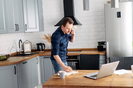 Solving urgent tasks. A young man speaks on the phone and prints a tect on a leptor on the go. Work from home, work remotelyの写真素材