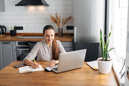 Online training. Girl student watching a lecture, webinar using a laptop. She sits in the kitchen at the table and writes information with a pen in a notebook.の写真素材