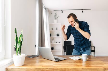 We did it! A young man in a casual shirt rejoices at job success, a successful deal, and profit growth. He is talking on the phone and looking at the laptop screen.の写真素材