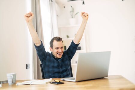 It's a success. A young guy in a checkered shirt sits in front of a laptop, from triumphantly raised his hands upの写真素材