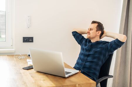 Rest at the workplace in the office. The young man in casual shirt sat back in a chair with satisfaction. A laptop on the tableの写真素材