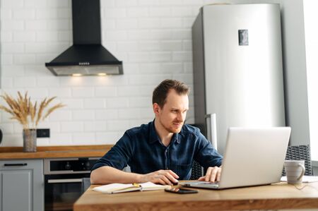 Young man in checkered shirt working on laptop in the kitchen at homeの写真素材