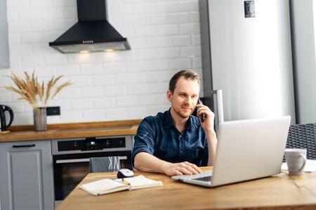 Freelancer in the kitchen at home. A young attractive guy uses a laptop for work, he sends emails and answers callsの写真素材
