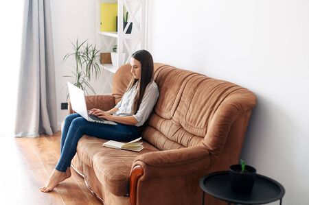 Closeup portrait of a young girl on a sofa at home. She is typing on a laptop and smiling. Chatting, online shopping, copywritingの写真素材