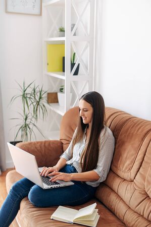 Closeup portrait of a young girl on a sofa at home. She is typing on a laptop and smiling. Chatting, online shopping, copywritingの写真素材