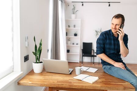 Office at home. A young man in a bright modern office space speaks on a mobile phone. On the table is a laptop, notebook for notes. Guy sits at the edge of the tableの写真素材