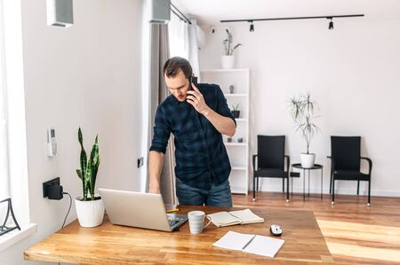 Office work. Young casual guy in a minimalistic modern office working using laptop. He is talking on a cell phone.の写真素材