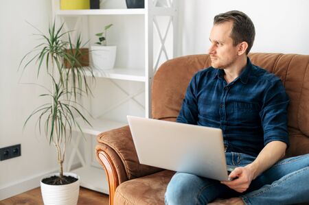 Closeup portrait of a young man with laptop in home interior. He is sitting on a sofa in a casual checkered shirt.の写真素材