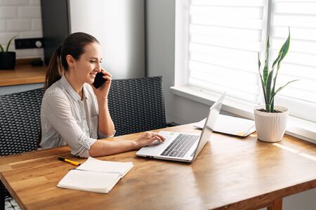 Home office, remote work, freelance. A young woman takes calls and uses a laptop for work, she sits in the kitchen in the houseの写真素材