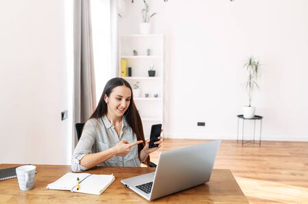 A female office employee is participating in an online conference. She points a finger on the screen of phone in a laptop camera.の写真素材