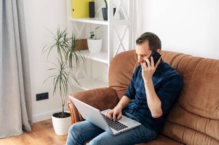 Home leisure. The guy is sitting at home on the sofa, talking on the phone and using a laptop. Online shopping, web surfing, social networksの写真素材