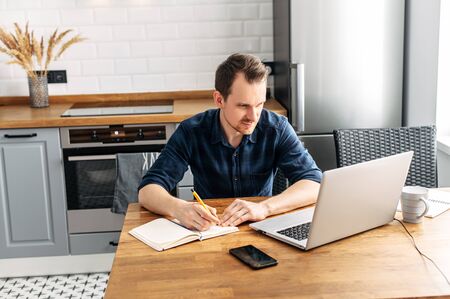 Home office, distant work, freelancer. A young man in a casual shirt in the kitchen at home uses a laptop. He writes the information with a pen in a notebook.の写真素材