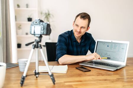 Portrait of a man with a laptop. He points to the laptop screen by pen, looks at the camera on a tripod. Recording video lessons, video blogsの写真素材