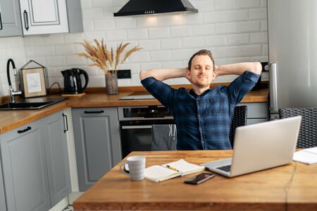 Home office, distant work, freelancer. Young man takes a break, resting leaning back in a chair in front of a laptop in the kitchen at homeの写真素材