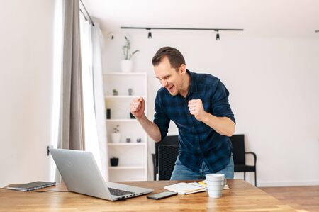 We did it! A young man in a casual shirt rejoices at job success, a successful deal, and profit growth. He looks at the laptop screen and triumphantly raised his hands.の写真素材