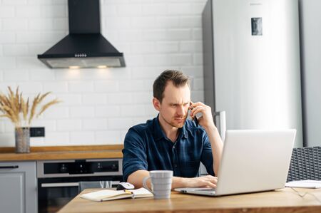 Freelancer in the kitchen at home. A young attractive guy uses a laptop for work, he sends emails and answers callsの写真素材