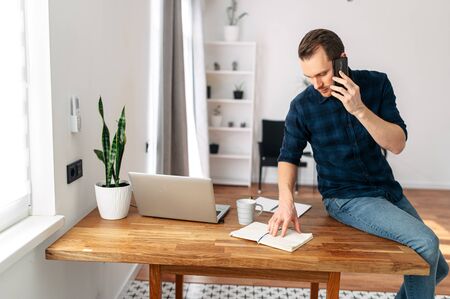 Office at home. A young man in a bright modern office space speaks on a mobile phone. On the table is a laptop, notebook for notes. Guy sits at the edge of the tableの写真素材