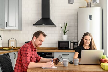 A married couple of freelancers work from home. A guy and a girl in casual clothes with gadgets work in the Scandinavian kitchen at the tableの写真素材