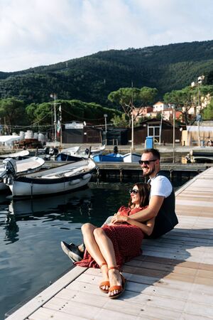 Love story on the pier. A handsome guy with a beard sits with his legs dangling, a beautiful girl in a red dress is lying on the guy. Yachts, boats and sea on the backgroundの写真素材