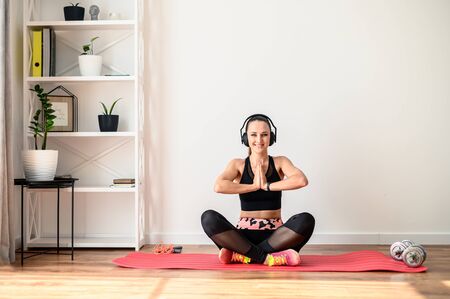 Positive and athletic girl is doing sport at home. She is sitting in a lotus position on the mat and looking at the camera and smiling, her hands are folded in greeting namaste, the headphones on herの写真素材