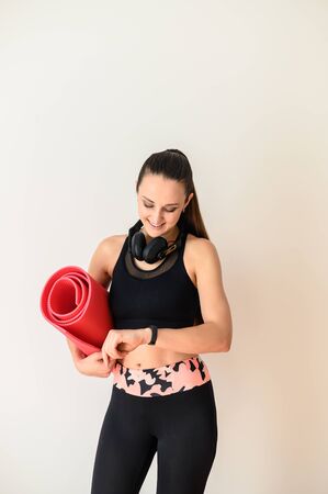 A slim, athletic girl in sportswear looks at a fitness tracker on her arm, koremat in her other hand. Sporty and healthy lifestyleの写真素材