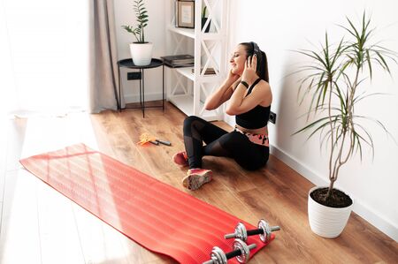 Doing sports at home, fitness at home. A young woman in a sports outfit sits and listens to music with headphones during a break. Koremat and sports equipment on the floorの写真素材
