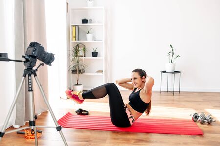 Video tutorial, online training. A young woman is recording a video home fitness lesson. She does fitness exercises on koremat, the camera on the tripod oppositeの写真素材