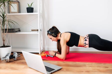 Video training in sports. A young woman does sports using a laptop to watch fitness tutorials at home, she does an exercise plankの写真素材