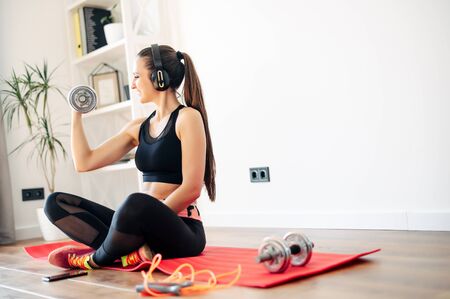 Sports at home during quarantine. Young woman in headphones sits on a mat and does exercises with dumbbellsの写真素材