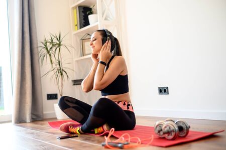 A positive and healthy girl in a sports outfit sits on a mat at home. She enjoys listening to music with headphones after training.の写真素材