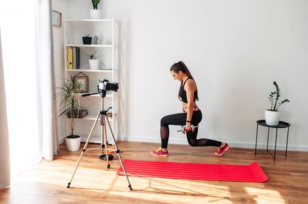 Video tutorial, online training. A young woman is recording a video home fitness lesson. She does fitness exercises lunges, the camera on the tripod oppositeの写真素材