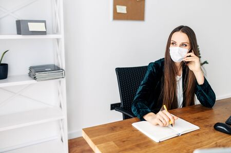 In touch in any situation. A woman in a smart casual works at the office during quarantine. She is in a medical mask talking on a smartphone and taking notes in a notebook.の写真素材