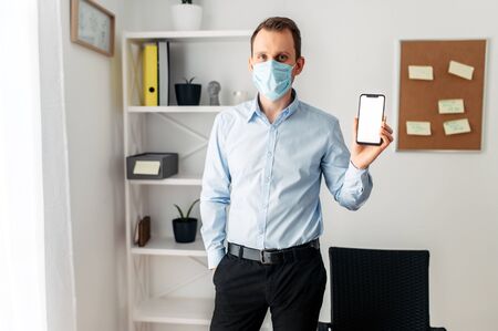 A guy in a formal shirt and a medical mask shows the screen of a smartphone. Work from home during the epidemic, stay homeの写真素材