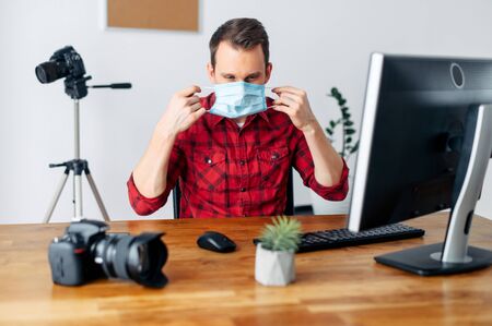 Freelancer photographer, videographer works using a medical mask from home during a pandemic, epidemic of coronavirus, seasonal flu. Young man uses computer for processing, photo equipment in the backgroundの写真素材