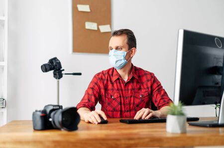 Freelancer photographer, videographer works using a medical mask from home during a pandemic, epidemic of coronavirus, seasonal flu. Young man uses computer for processing, photo equipment in the backgroundの写真素材