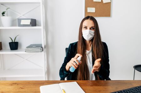 Protect yourself during a pandemic, epidemic. Young woman in office spraying a disinfector on her hands, a medical mask on her. Front viewの写真素材