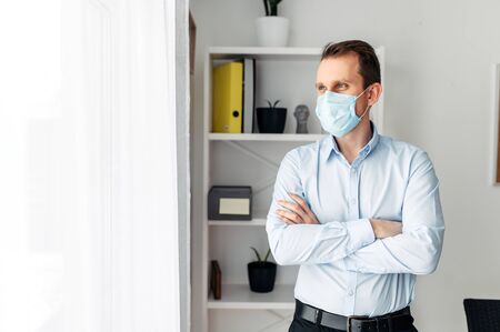 Protect yourself, be careful. A young man in an office in a medical mask stands with his arms crossed on his chest. Pandemic, seasonal diseasesの写真素材