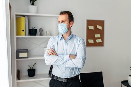 Protect yourself, be careful. A young man in an office in a medical mask stands with his arms crossed on his chest. Pandemic, seasonal diseasesの写真素材