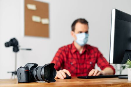 Stay at home. Work from home, office at home. A young guy, a videographer photographer in a medical mask, processes material at home on a computer, his face is out of focus. Coronavirus pandemicの写真素材