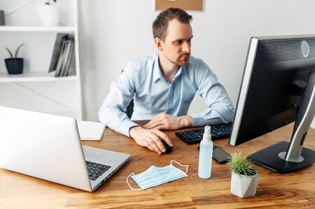 The man stayed at home and works remotely. The guy uses a computer and a laptop for work, and medicine mask and antiseptic for self protective. Coronavirus pandemic, influenza, covid 19の写真素材