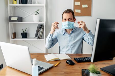 A young man fulfills recommendations to prevent the spread of the virus in the office. He is putting on a protective medical mask in front of a computerの写真素材