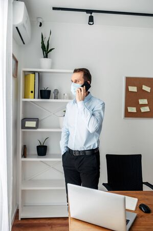 Stay at home. A young man works from home, home office, work remotely. Medical masked businessman talking on cell phoneの写真素材