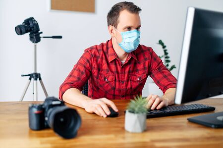 Freelancer photographer, videographer works using a medical mask from home during a pandemic, epidemic of coronavirus, seasonal flu. Young man uses computer for processing, photo equipment in the backgroundの写真素材